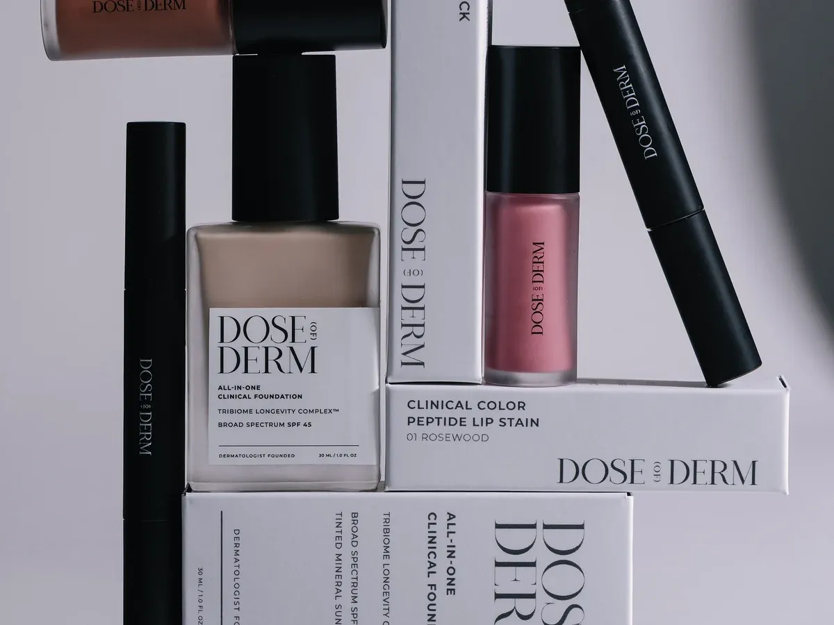 DOSE (OF) DERM All-in-One Clinical Foundation SPF 45 in a 30 ml bottle, alongside a Clinical Color Peptide Lip Stain carton labelled 01 Rosewood and black dual sticks; foundation shade not specified.