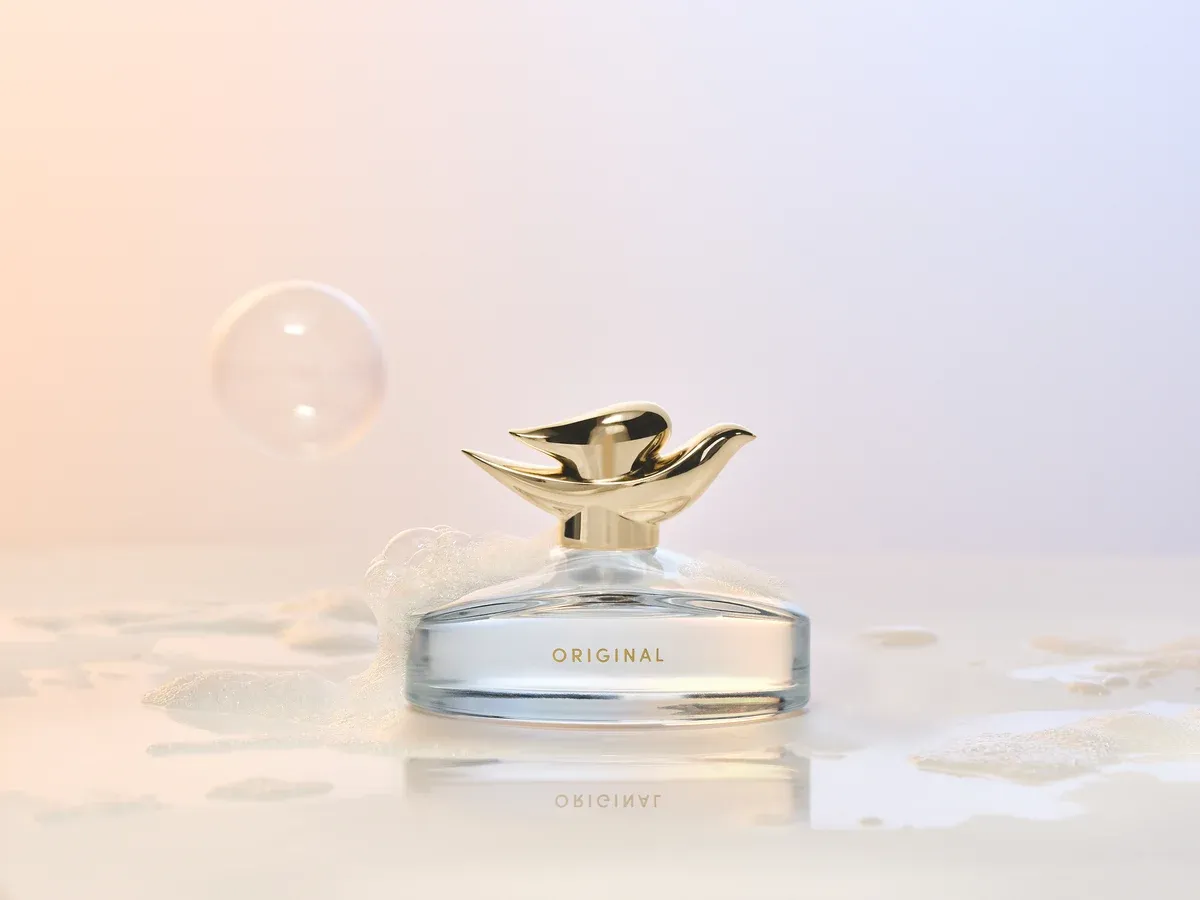 Dove Original Fragrance in a clear, curved glass bottle marked Original, with a gold dove-shaped cap, surrounded by bubbles and suds on a peach-and-lilac background.