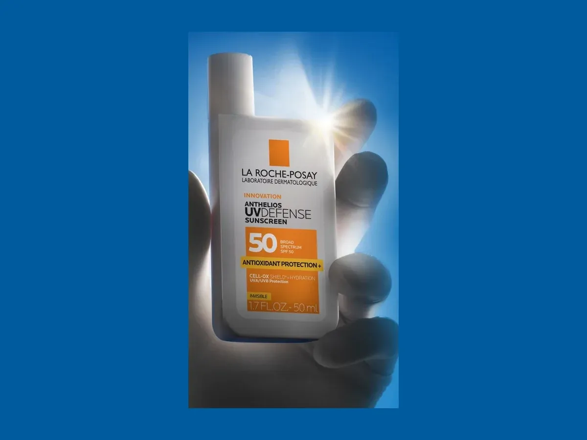 Close-up of a hand holding the white La Roche-Posay Anthelios UVDefense Sunscreen SPF 50 bottle with BEMT, US 50 mL / 1.7 fl oz version, against blue light.