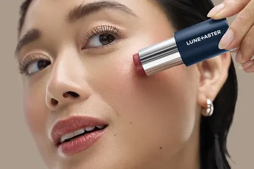 Model applying Lune+Aster Cream Multi-Stick from its navy-and-silver tube to her cheek; rosy shade not identified in the brand’s press caption.
